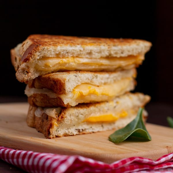 Stack of three grilled cheese sandwiches with melted cheese on a wooden board with a red checkered napkin.