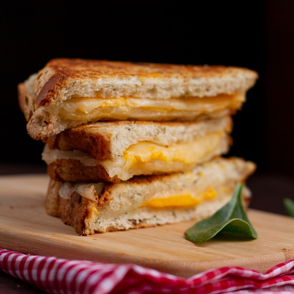 Stack of three grilled cheese sandwiches with melted cheese on a wooden board with a red checkered napkin.