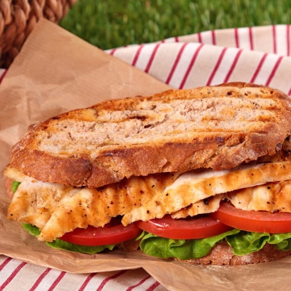 Grilled chicken sandwich with lettuce and tomato on toasted bread, wrapped in paper on a striped cloth.