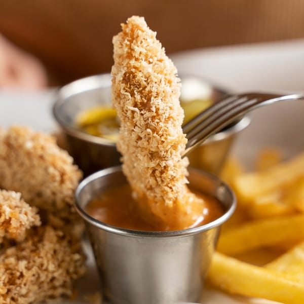 Crispy chicken tender being dipped into a sauce cup, with fries and more tenders in the background.